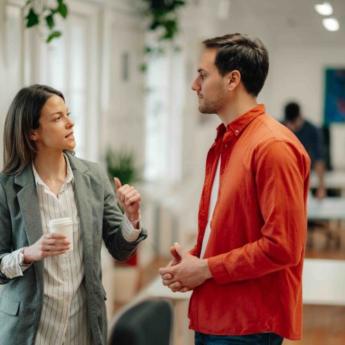 Businesswoman holding a disposable coffee cup, engaging in an informal conversation with a businessman in an office hallway, discussing work related topics and ideas